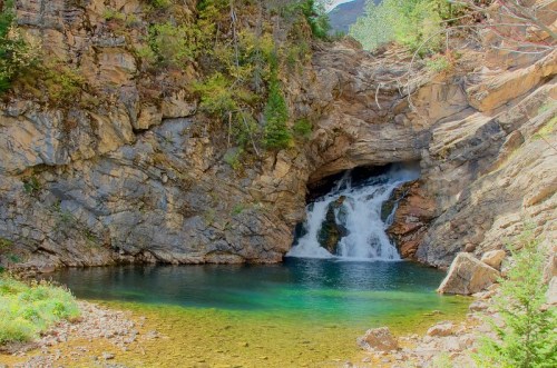 A serene waterfall cascades into a clear pool, surrounded by rocky cliffs and lush greenery.