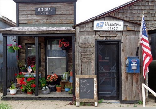 A rustic general store with flower pots, a mailbox, and an American flag, showcasing a charming small-town vibe.