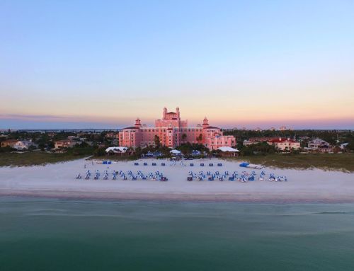 Aerial view of a pink hotel by the beach at sunset, with blue lounge chairs arranged on the sandy shore.