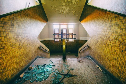 Abandoned staircase with broken glass, peeling walls, and a dimly lit landing at the top.