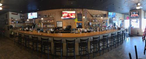 A panoramic view of a bar with a long counter, shelves of drinks, and TVs displaying sports.