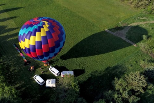 A colorful hot air balloon on a grassy field, casting a large shadow, with two vans nearby.