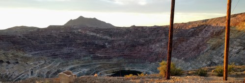 A panoramic view of a large open-pit mine with layered rock formations and a distant mountain under a sunset sky.