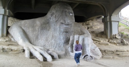 A person stands near a large, whimsical troll sculpture under a bridge, with its massive hands and face visible.