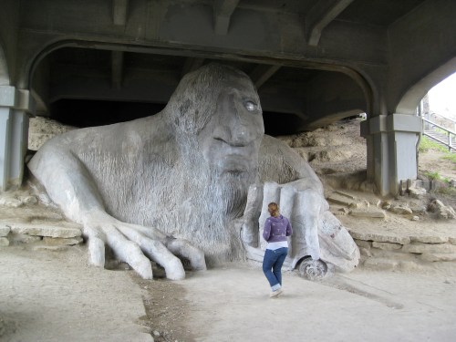 A large concrete troll sculpture under a bridge, with a person walking nearby, showcasing its massive size and detail.
