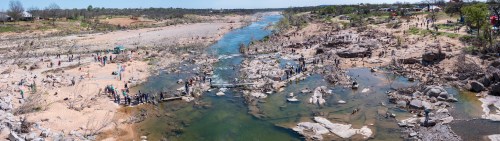 A panoramic view of a riverbank with people exploring rocky areas and a flowing river under a clear sky.