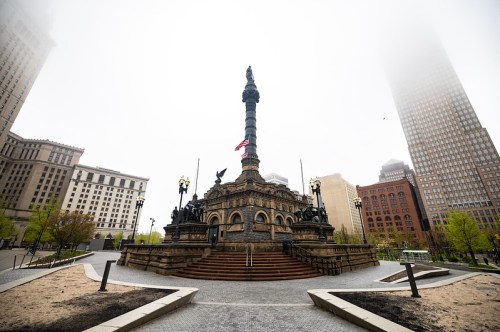 A foggy view of a tall monument surrounded by buildings and greenery in an urban park setting.