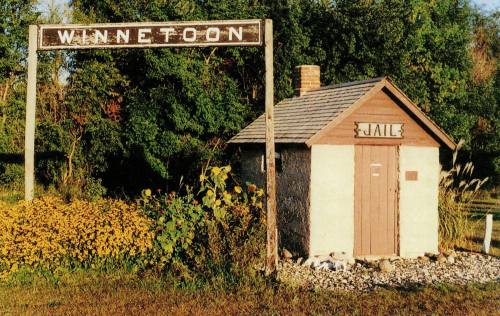 A small, rustic jail building with a sign reading "Winnetoon" above it, surrounded by greenery and colorful foliage.