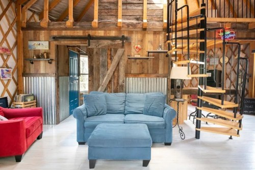 Cozy interior of a rustic cabin featuring a blue couch, red chair, and a spiral staircase in a wooden setting.