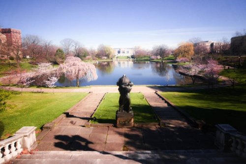 A serene park scene featuring a statue by a pond surrounded by blooming trees and clear blue skies.