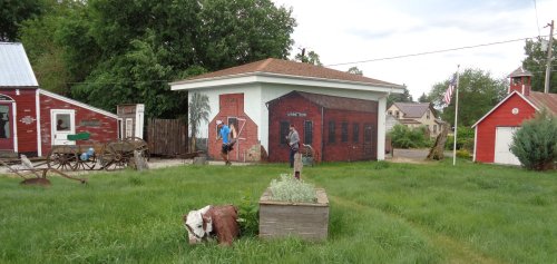 A grassy area with a mural of a red barn on a building, surrounded by small structures and a flagpole in the background.