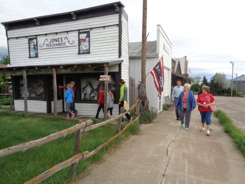 A group of people walks past a historic building with a sign reading "Jones Merchandise" and a flag nearby.