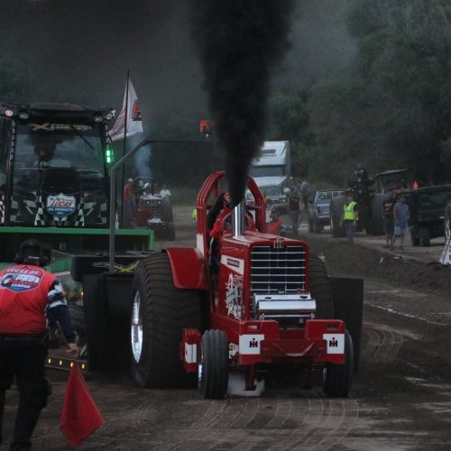 A red tractor pulling vehicle emits black smoke during a competition, with spectators and other vehicles in the background.
