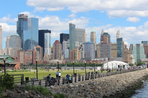 A skyline view of tall buildings and skyscrapers along a waterfront, with people walking and green spaces in the foreground.
