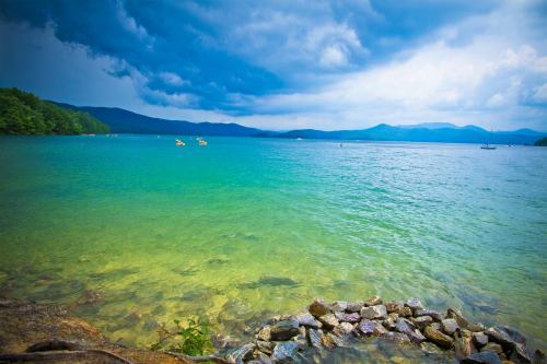 A serene lake with clear turquoise water, rocky shore, and dark clouds overhead, surrounded by green hills.