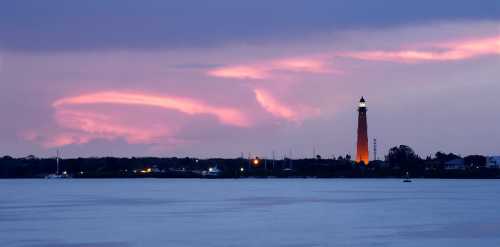 A lighthouse stands by the water at dusk, illuminated against a colorful sky with clouds and distant lights.