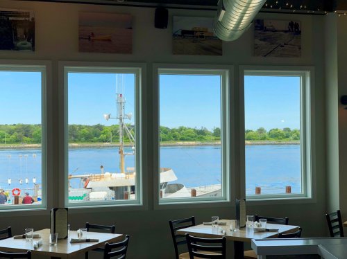 View of a serene waterfront through large windows, with boats and greenery visible outside. Bright and inviting atmosphere.
