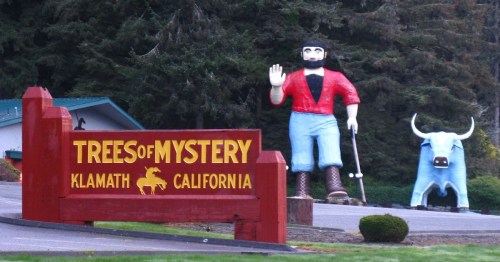 A large statue of a lumberjack waves next to a blue bull, with a sign reading "Trees of Mystery" in Klamath, California.