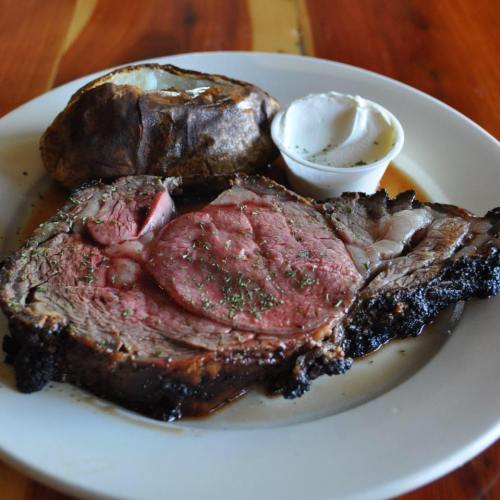 A plate featuring a slice of juicy prime rib with herbs, a baked potato, and a side of sour cream.