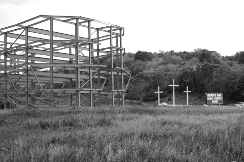 A skeletal metal structure beside three large crosses and a sign reading "Noah's Ark" in a grassy field.