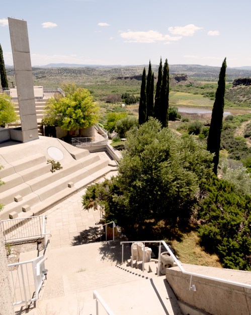 A modern architectural structure with steps, greenery, and tall trees overlooking a river and distant hills under a clear sky.