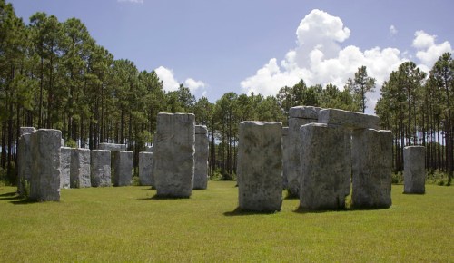 A stone monument resembling Stonehenge, surrounded by grass and tall trees under a blue sky with clouds.