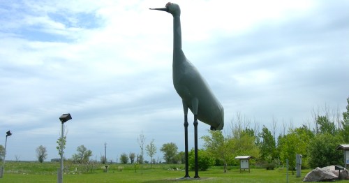 A large metal sculpture of a crane stands in a grassy area, surrounded by trees and a cloudy sky.