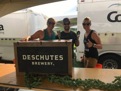 Three people stand behind a wooden bar with "Deschutes Brewery" signage, smiling and holding beer taps at an outdoor event.
