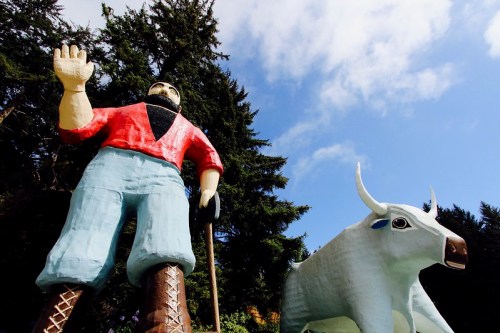 A large statue of a man in a red shirt and blue pants stands next to a white cow under a blue sky and trees.