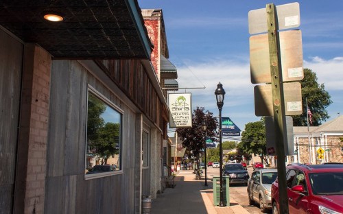 A street view featuring shops, signage, and parked cars under a clear blue sky.