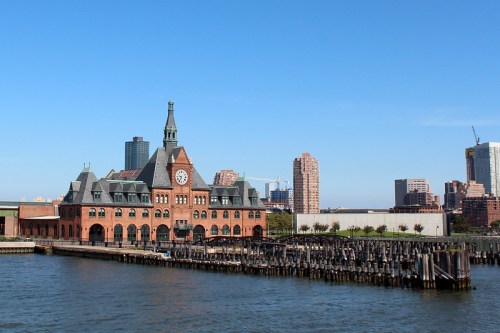 Historic brick building with a clock tower by the water, surrounded by modern city skyline and clear blue sky.