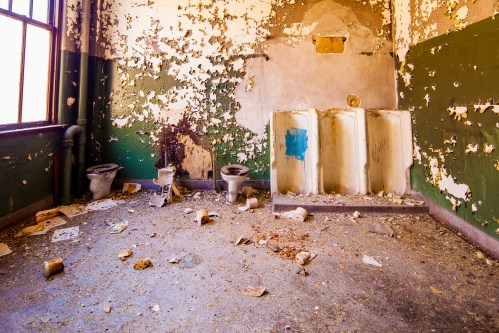 Abandoned restroom with peeling paint, two toilets, and a urinal, littered with debris on the floor.