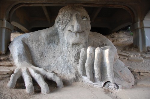 A large concrete troll sculpture with a menacing expression, sitting under a bridge, holding a toy car.