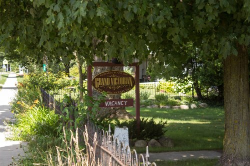 Sign for "Grand Victorian" with "Vacancy" under a leafy tree, surrounded by a well-kept garden and sidewalk.