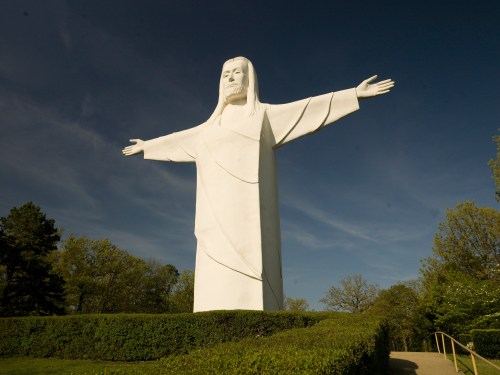 A large white statue of Christ with outstretched arms, surrounded by greenery under a clear blue sky.
