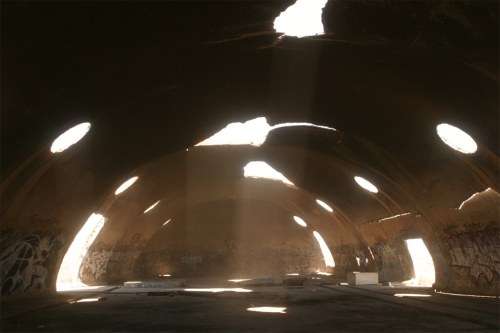 Interior of a dilapidated building with an arched ceiling, sunlight streaming through circular openings, and graffiti on the walls.