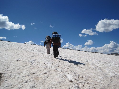 Two hikers with backpacks walk on a snowy landscape under a bright blue sky with fluffy clouds.