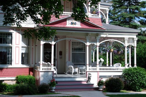 A charming Victorian-style house with a wraparound porch, white rocking chairs, and colorful flower beds.