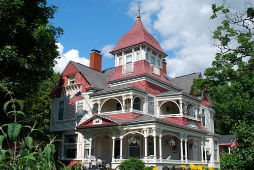 A large, ornate Victorian-style house with a red roof, intricate details, and a wraparound porch surrounded by greenery.