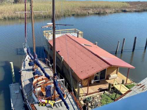 Aerial view of a boat docked next to a wooden house on stilts by a calm waterway, surrounded by tall grass.