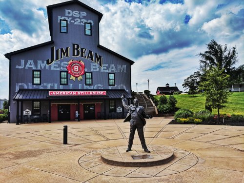 A statue of a man in front of the Jim Beam distillery, with a large building and blue sky in the background.