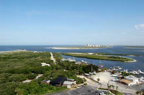 Aerial view of a coastal area with boats, lush greenery, and a sandy shoreline under a clear blue sky.