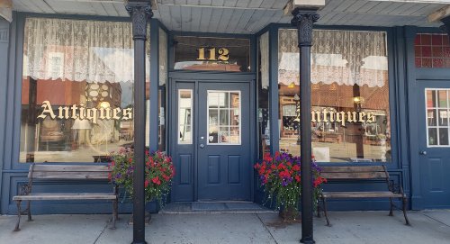 A charming antique shop with large windows, floral displays, and benches outside, located at 112 Main Street.