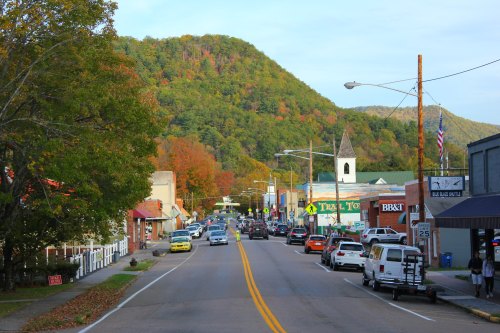 A small town street lined with shops and trees, with a mountain backdrop and autumn foliage.