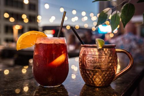 Two cocktails on a table: a fruity drink with an orange slice and a copper mug with a lime wedge, with string lights in the background.