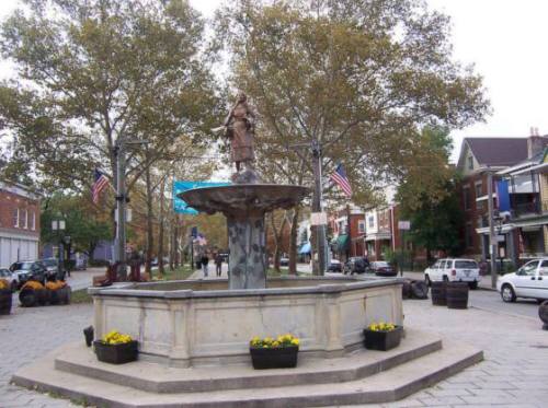 A stone fountain with a statue in a park, surrounded by trees, planters, and buildings, with people walking nearby.