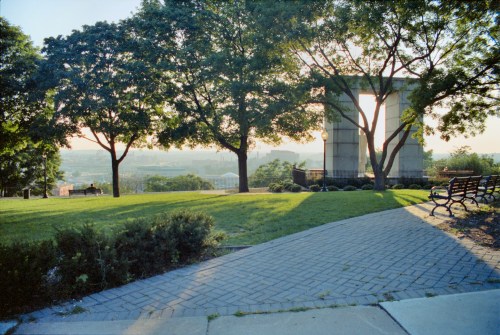 A park scene with trees, a stone monument, and a pathway leading to a view of the landscape in the background.