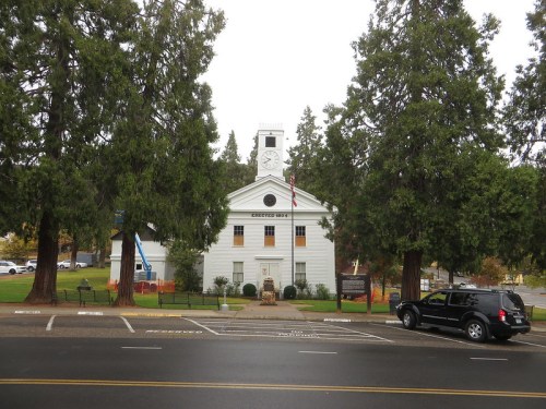 A white building with a clock tower, surrounded by tall trees, on a rainy day. A black SUV is parked nearby.