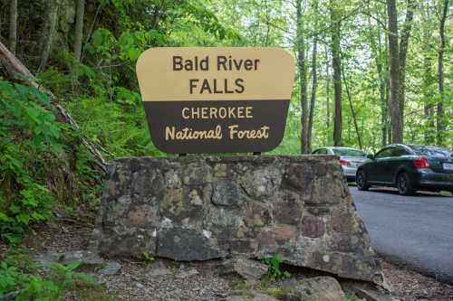 Sign for Bald River Falls in Cherokee National Forest, surrounded by lush greenery and parked cars nearby.