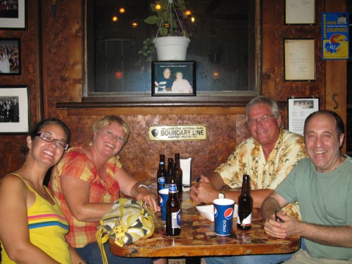 A group of four friends sitting at a table in a cozy bar, enjoying drinks and snacks, smiling at the camera.